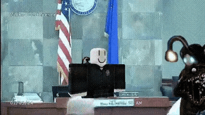 A Roblox Character Is Sitting At A Desk In Front Of A Computer In A Courtroom With An American Flag In The Background . GIF