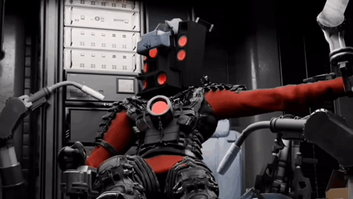 A Robot With A Red Head Is Sitting In A Chair In Front Of A Computer GIF