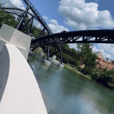 A Roller Coaster Going Over A Body Of Water With Trees In The Background GIF