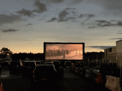 A Row Of Cars Are Parked In Front Of A Large Screen That Shows A Movie GIF