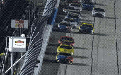 A Row Of Race Cars Are Driving Down A Track With A Sign That Says Cook Out GIF