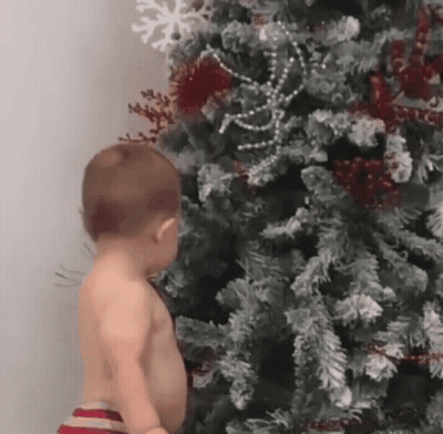 A Shirtless Baby Is Standing Next To A Christmas Tree . GIF
