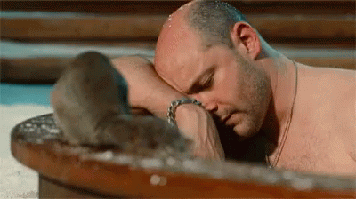 A Shirtless Bald Man Is Laying On A Wooden Table With His Head On His Arm . GIF