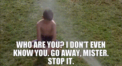 A Shirtless Boy Is Sitting In The Grass With A Sprinkler Spraying Water On Him . GIF