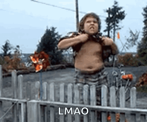 A Shirtless Boy Is Standing In Front Of A Fence And Holding His Chest . GIF