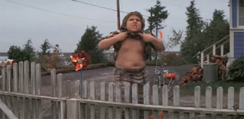A Shirtless Boy Is Standing In Front Of A Wooden Fence With A Pinwheel In The Background . GIF