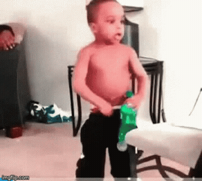 A Shirtless Little Boy Is Standing In A Room Holding A Bottle Of Soda . GIF