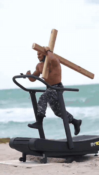 A Shirtless Man Carrying A Wooden Cross On A Treadmill On The Beach GIF