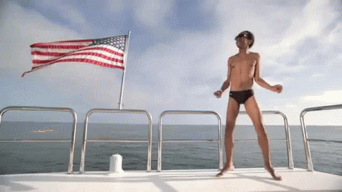 A Shirtless Man Dancing On A Boat In Front Of An American Flag GIF