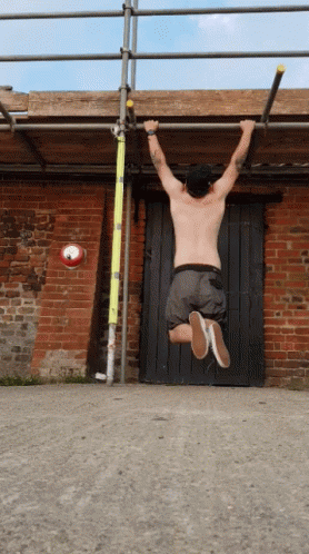 A Shirtless Man Hangs Upside Down On A Bar In Front Of A Brick Wall GIF