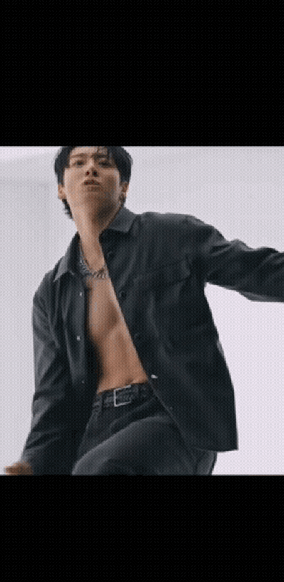 A Shirtless Man In A Black Jacket Is Standing In Front Of A White Wall With His Arms Outstretched . GIF