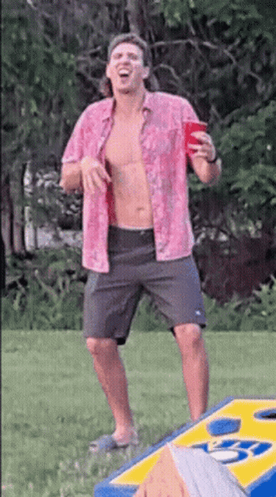 A Shirtless Man In A Pink Shirt Is Holding A Red Cup While Standing Next To A Cornhole Board . GIF