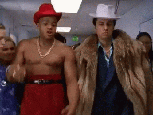 A Shirtless Man In A Red Cowboy Hat And A Fur Coat Is Walking Down A Hallway With A Group Of People . GIF
