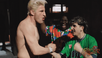 A Shirtless Man In A Street Fighter Shirt Holds Hands With Another Shirtless Man GIF