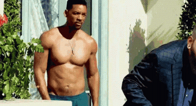 A Shirtless Man In Blue Shorts Is Standing Next To Another Man In A Suit . GIF
