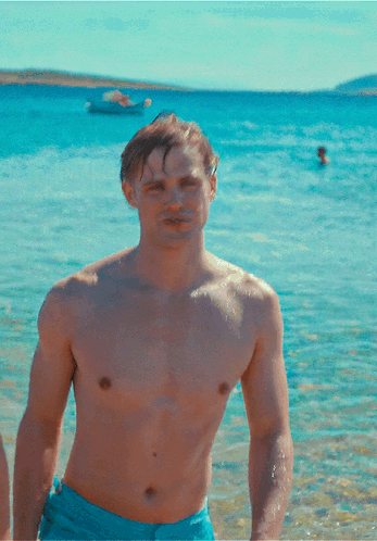 A Shirtless Man In Blue Shorts Stands On A Beach GIF