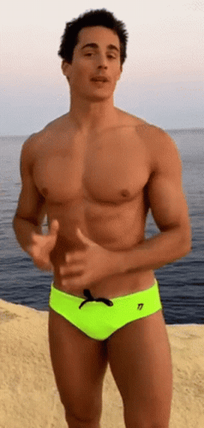 A Shirtless Man In Neon Yellow Swim Trunks Is Standing On The Beach Near The Ocean . GIF