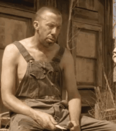 A Shirtless Man In Overalls Is Sitting In Front Of A Window GIF