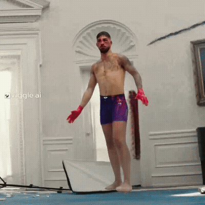 A Shirtless Man In Purple Shorts And Red Gloves Stands On A Blue Mat In A Room GIF