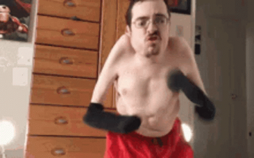 A Shirtless Man In Red Shorts And Black Gloves Is Standing In Front Of A Dresser . GIF
