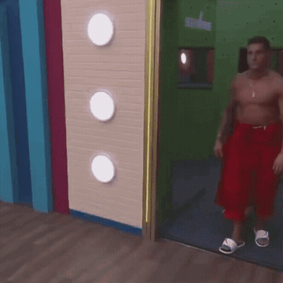 A Shirtless Man In Red Shorts And White Sandals Is Walking Through A Door GIF