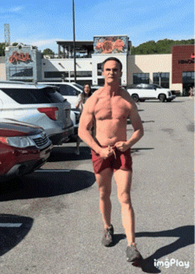 A Shirtless Man In Red Shorts Is Standing In A Parking Lot In Front Of A Building That Says King Crab GIF