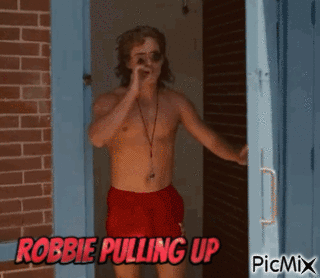 A Shirtless Man In Red Shorts Is Standing In Front Of A Brick Wall With The Words Robbie Pulling Up On The Bottom GIF