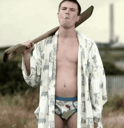 A Shirtless Man In Underwear Is Smoking A Cigarette While Holding A Large Wooden Stick Over His Shoulder . GIF