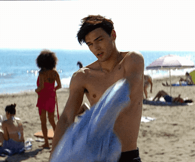 A Shirtless Man Is Holding A Blue Towel On A Beach GIF