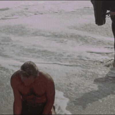 A Shirtless Man Is Kneeling On The Beach Near The Ocean GIF
