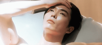 A Shirtless Man Is Laying In A Bathtub With His Hand On His Forehead GIF