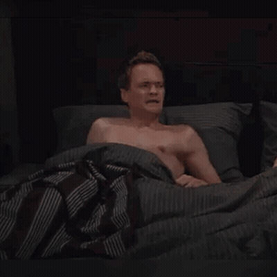 A Shirtless Man Is Laying In Bed With The Words Whaaat ? Written On The Bottom Of The Image . GIF