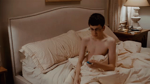 A Shirtless Man Is Laying On A Bed Looking At His Cell Phone GIF