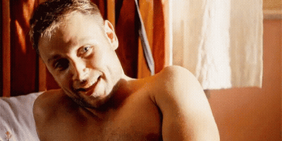 A Shirtless Man Is Laying On A Bed Smiling At The Camera . GIF