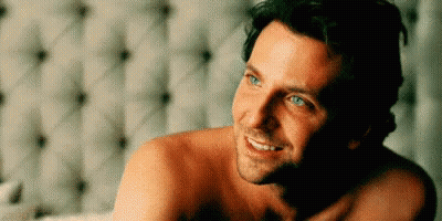 A Shirtless Man Is Laying On A Bed Smiling . GIF