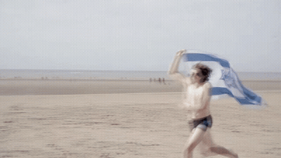 A Shirtless Man Is Running On The Beach Holding A Flag With A Blue And White Star On It GIF