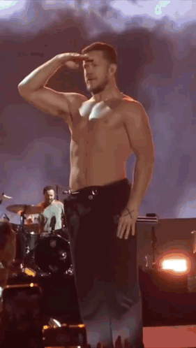 A Shirtless Man Is Saluting On A Stage In Front Of A Drummer . GIF