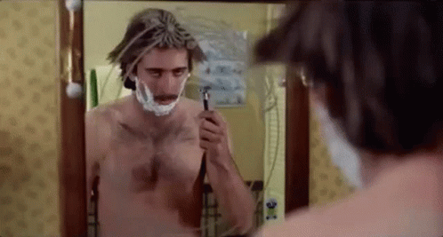A Shirtless Man Is Shaving His Face In Front Of A Bathroom Mirror . GIF