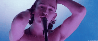 A Shirtless Man Is Singing Into A Microphone While Holding His Head . GIF