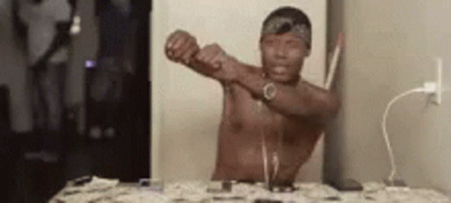 A Shirtless Man Is Sitting At A Table With His Arms Outstretched In Front Of A Television . GIF