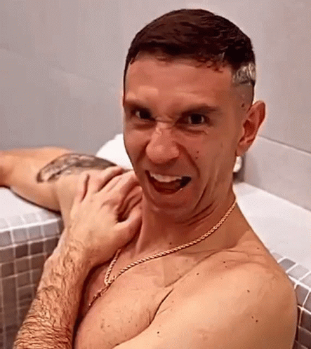A Shirtless Man Is Sitting In A Bathtub Making A Funny Face While A Woman Holds His Arm . GIF
