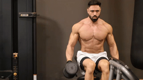 A Shirtless Man Is Sitting On A Bench Holding A Dumbbell With The Number 10 On It GIF