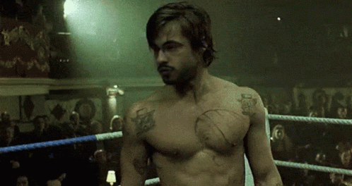 A Shirtless Man Is Standing In A Boxing Ring With A Crowd Watching . GIF