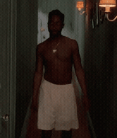 A Shirtless Man Is Standing In A Hallway Wearing White Shorts And A Necklace . GIF