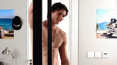 A Shirtless Man Is Standing In Front Of A Door With A Picture Of A Motorcycle On The Wall Behind Him . GIF