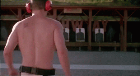 A Shirtless Man Is Standing In Front Of A Shooting Range Wearing Ear Defenders . GIF