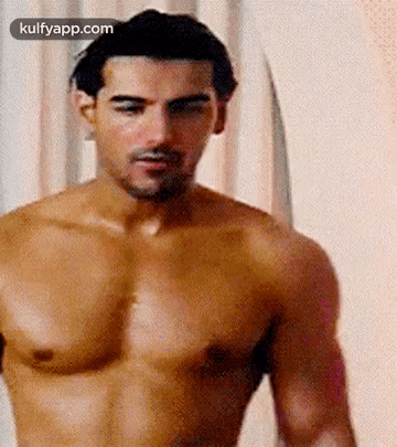 A Shirtless Man Is Standing In Front Of A White Curtain And Looking At The Camera . GIF
