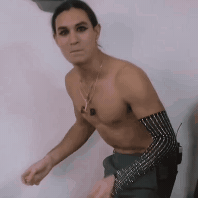 A Shirtless Man Is Standing In Front Of A White Wall Wearing A Necklace And A Bracelet . GIF