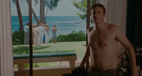 A Shirtless Man Is Standing In Front Of A Window Looking Out At The Ocean . GIF