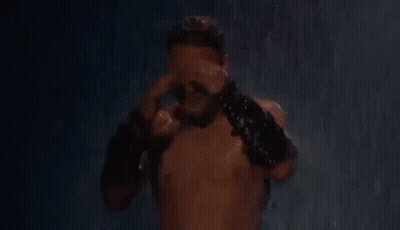 A Shirtless Man Is Standing In The Rain Holding His Head And Wearing Leather Gloves . GIF